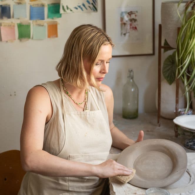 A woman in an apron shapes a clay bowl in a pottery studio with plants and art supplies surrounding her.