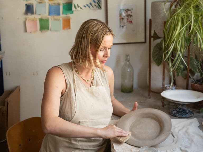 A woman in an apron shapes a clay bowl in a pottery studio with plants and art supplies surrounding her.