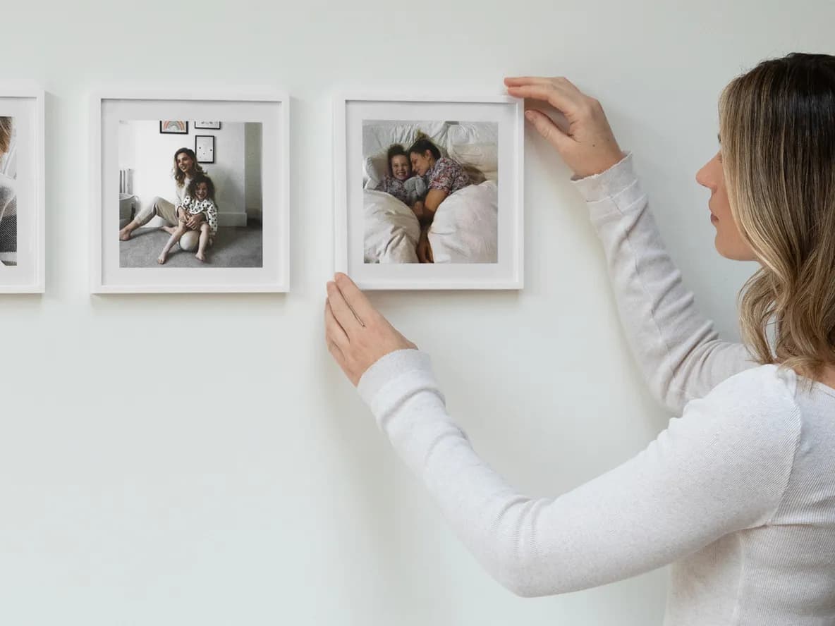 A woman hangs a framed photo of a mother and child sleeping on a white wall, completing a gallery of family moments.