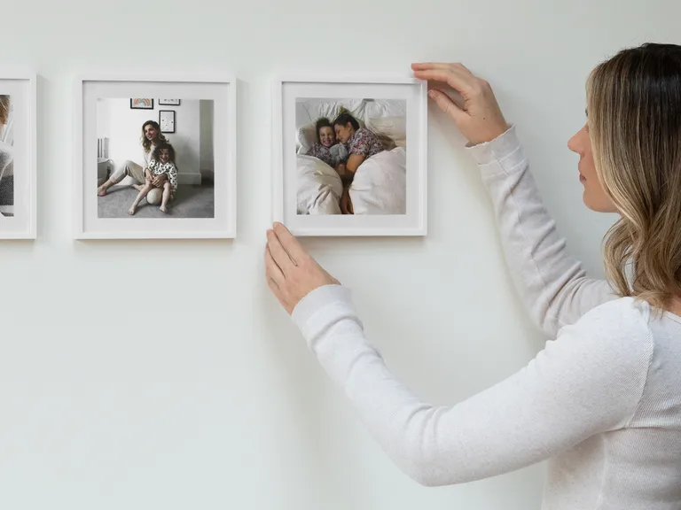 A woman hangs a framed photo of a mother and child sleeping on a white wall, completing a gallery of family moments.