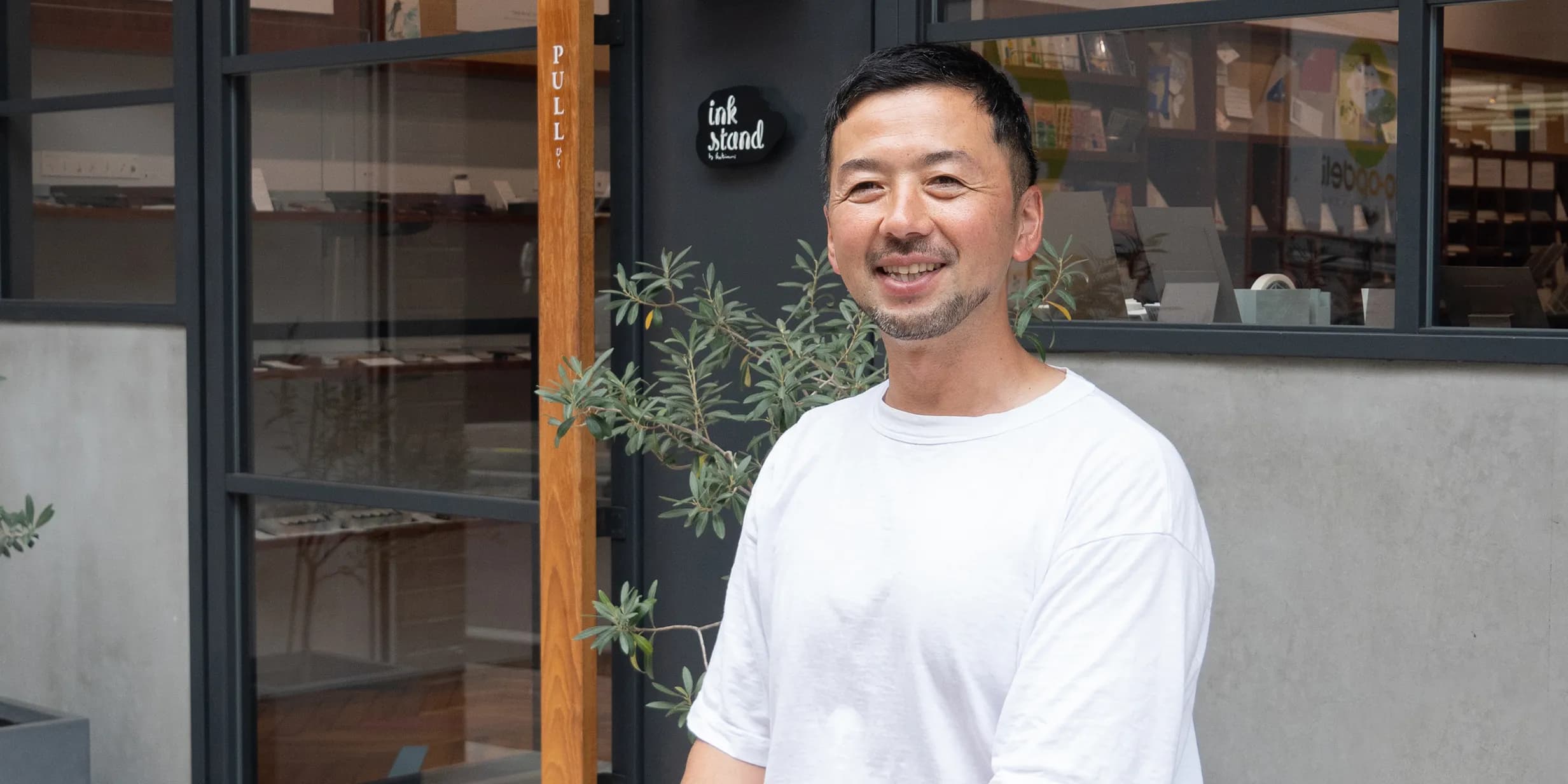 A smiling man in a white t-shirt stands outside a shop named "ink stand," with plants flanking the entrance and glass windows reflecting the interior.
