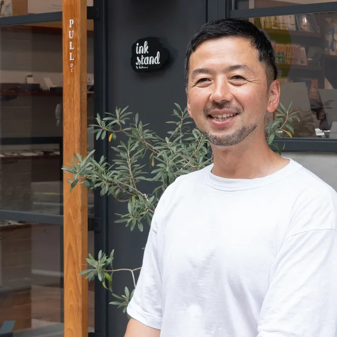 A smiling man in a white t-shirt stands outside a shop named "ink stand," with plants flanking the entrance and glass windows reflecting the interior.