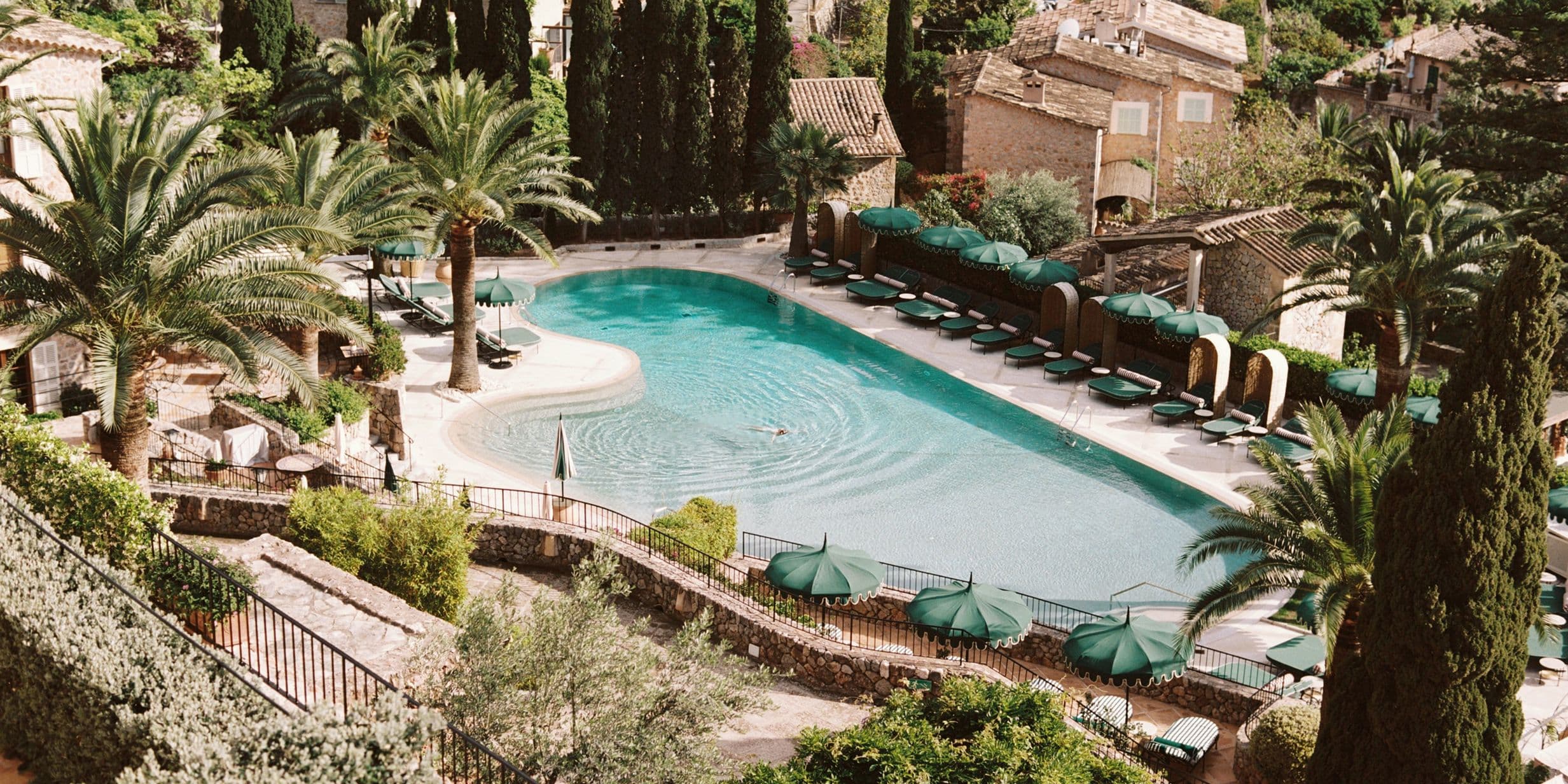 Aerial view of a serene pool surrounded by palm trees and sun loungers, with rustic stone buildings in the background.