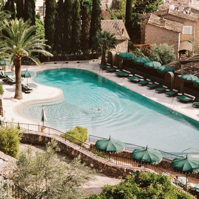 Aerial view of a serene pool surrounded by palm trees and sun loungers, with rustic stone buildings in the background.
