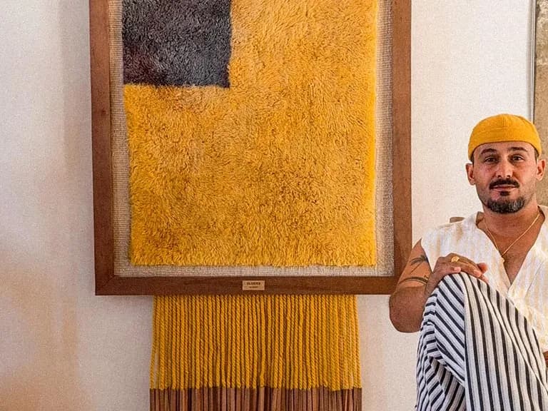 A man in a yellow hat sits by a vibrant yellow and black textured artwork, with a clay vase holding dried branches nearby in a rustic room.
