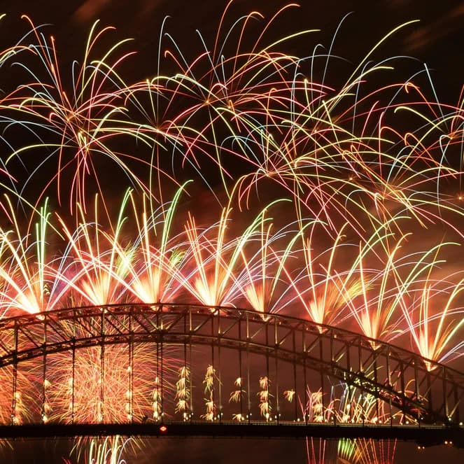Colorful fireworks light up the night sky over a large, arched bridge silhouetted against glowing bursts.
