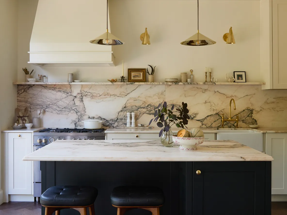 Elegant kitchen with marble countertops, white cabinets, black island, pendant lights, and large window. Decor includes plant and small frames.
