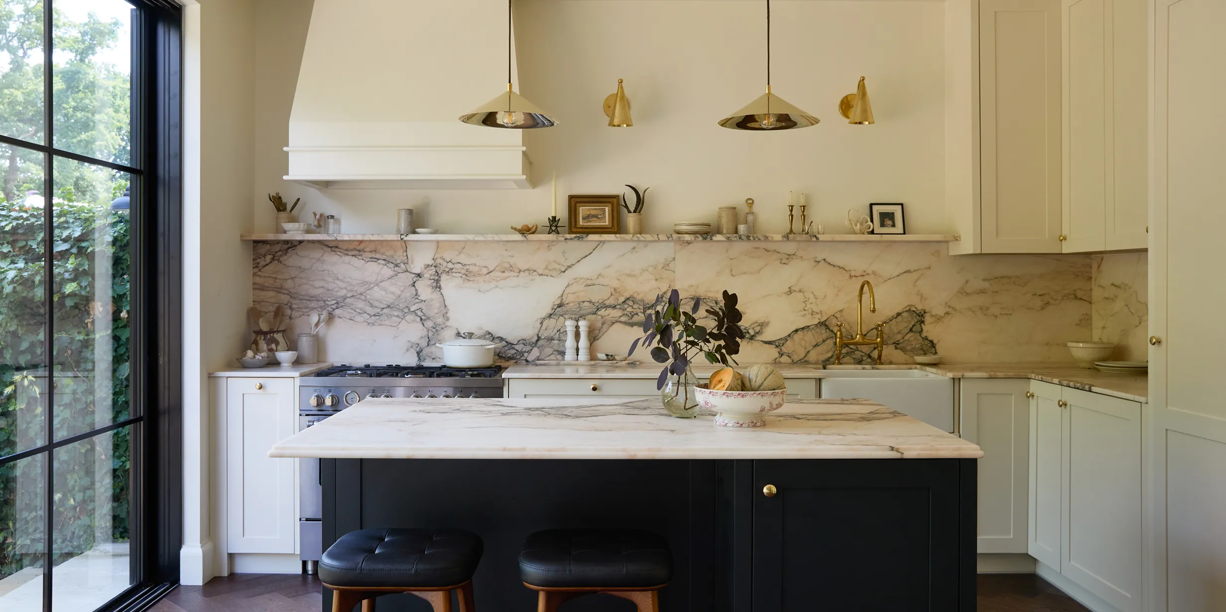 Elegant kitchen with marble countertops, white cabinets, black island, pendant lights, and large window. Decor includes plant and small frames.