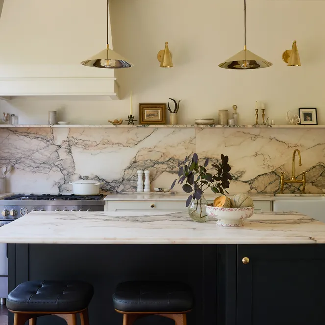 Elegant kitchen with marble countertops, white cabinets, black island, pendant lights, and large window. Decor includes plant and small frames.