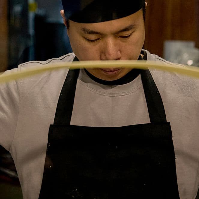 A chef in a black apron and hat stretches dough.