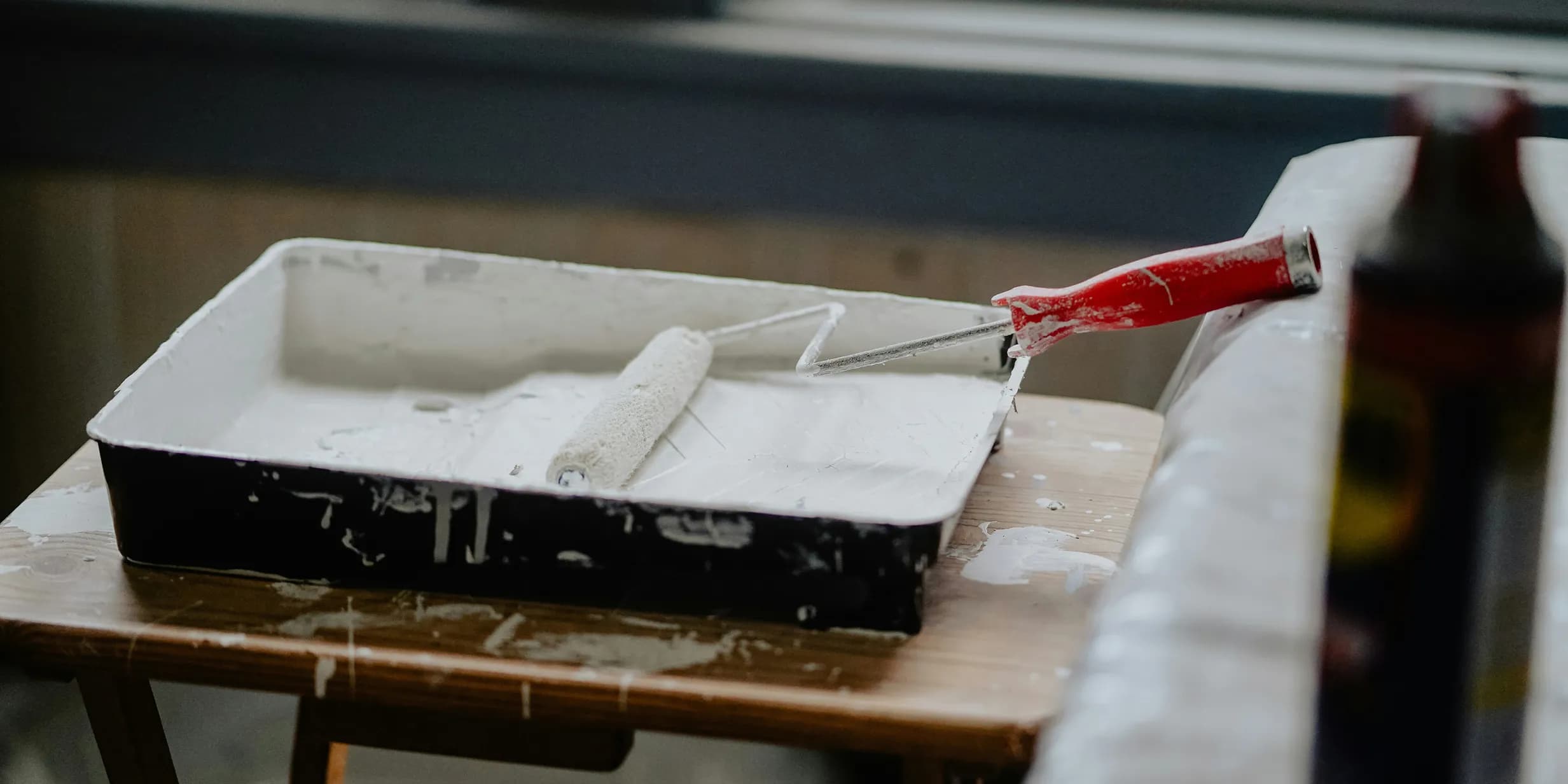 A paint roller with a red handle rests in a tray filled with white paint, placed on a wooden table.