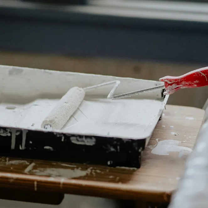 A paint roller with a red handle rests in a tray filled with white paint, placed on a wooden table.