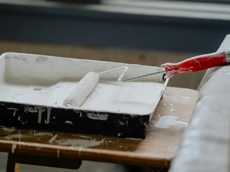 A paint roller with a red handle rests in a tray filled with white paint, placed on a wooden table.