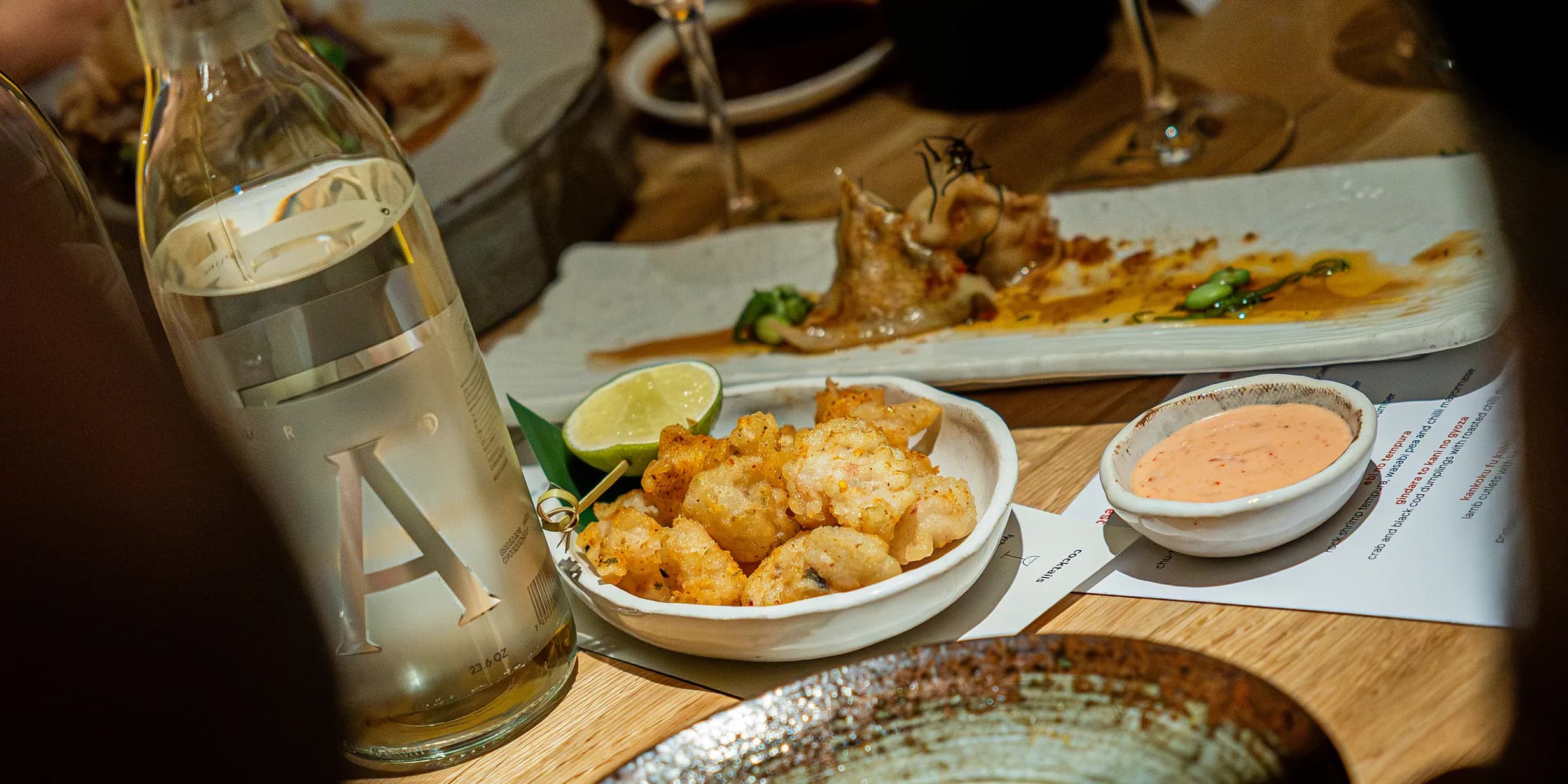 A table setting with a bottle of water, a plate of fried food with lime, dipping sauce, and a partially eaten dish in the background.