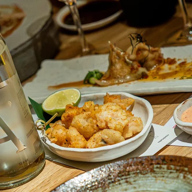 A table setting with a bottle of water, a plate of fried food with lime, dipping sauce, and a partially eaten dish in the background.