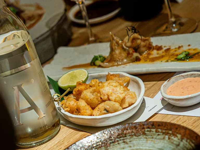 A table setting with a bottle of water, a plate of fried food with lime, dipping sauce, and a partially eaten dish in the background.