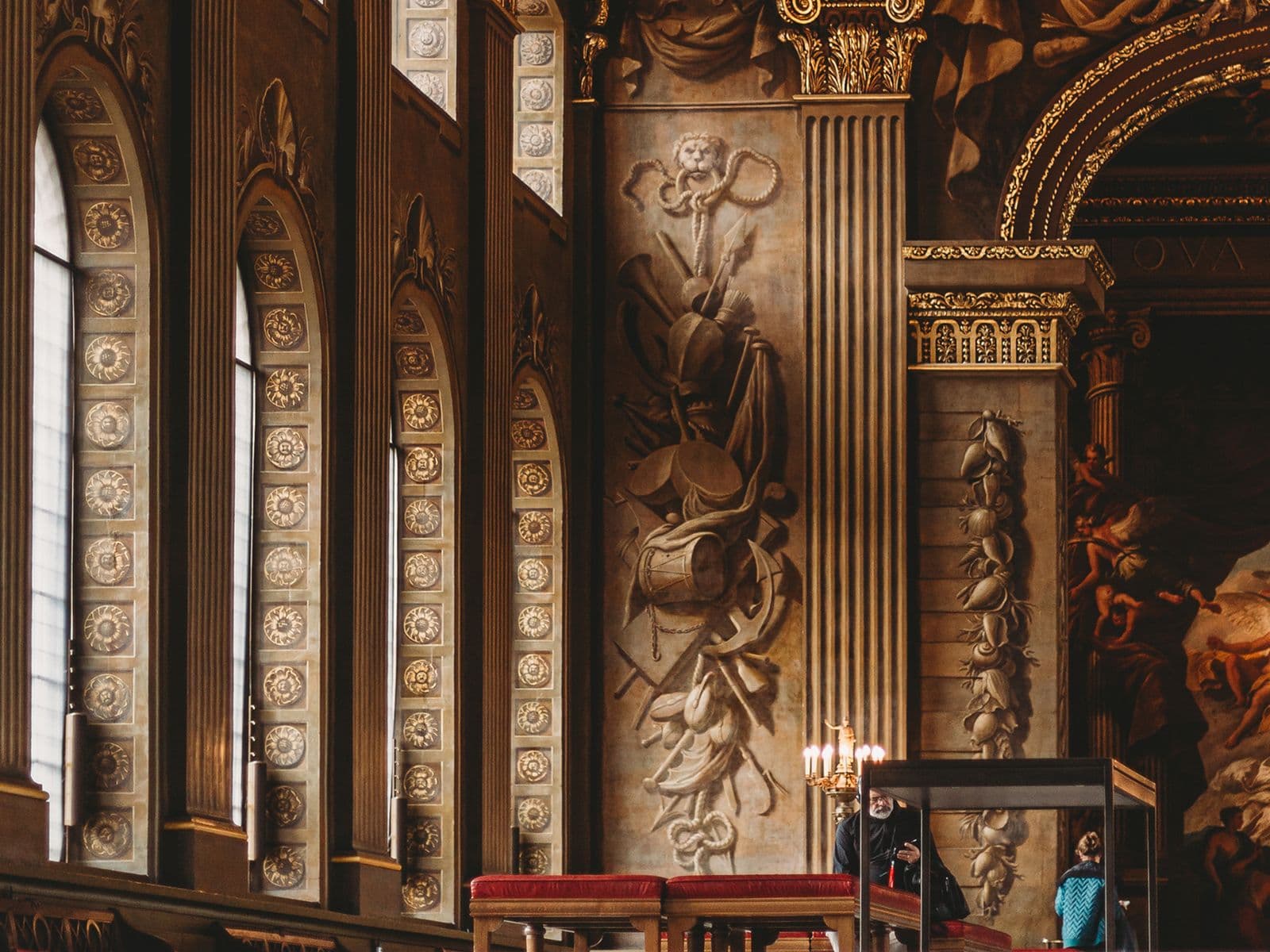 Ornate interior of a grand hall with tall arched windows, intricate carvings, and a detailed mural. Warm lighting adds a majestic feel.