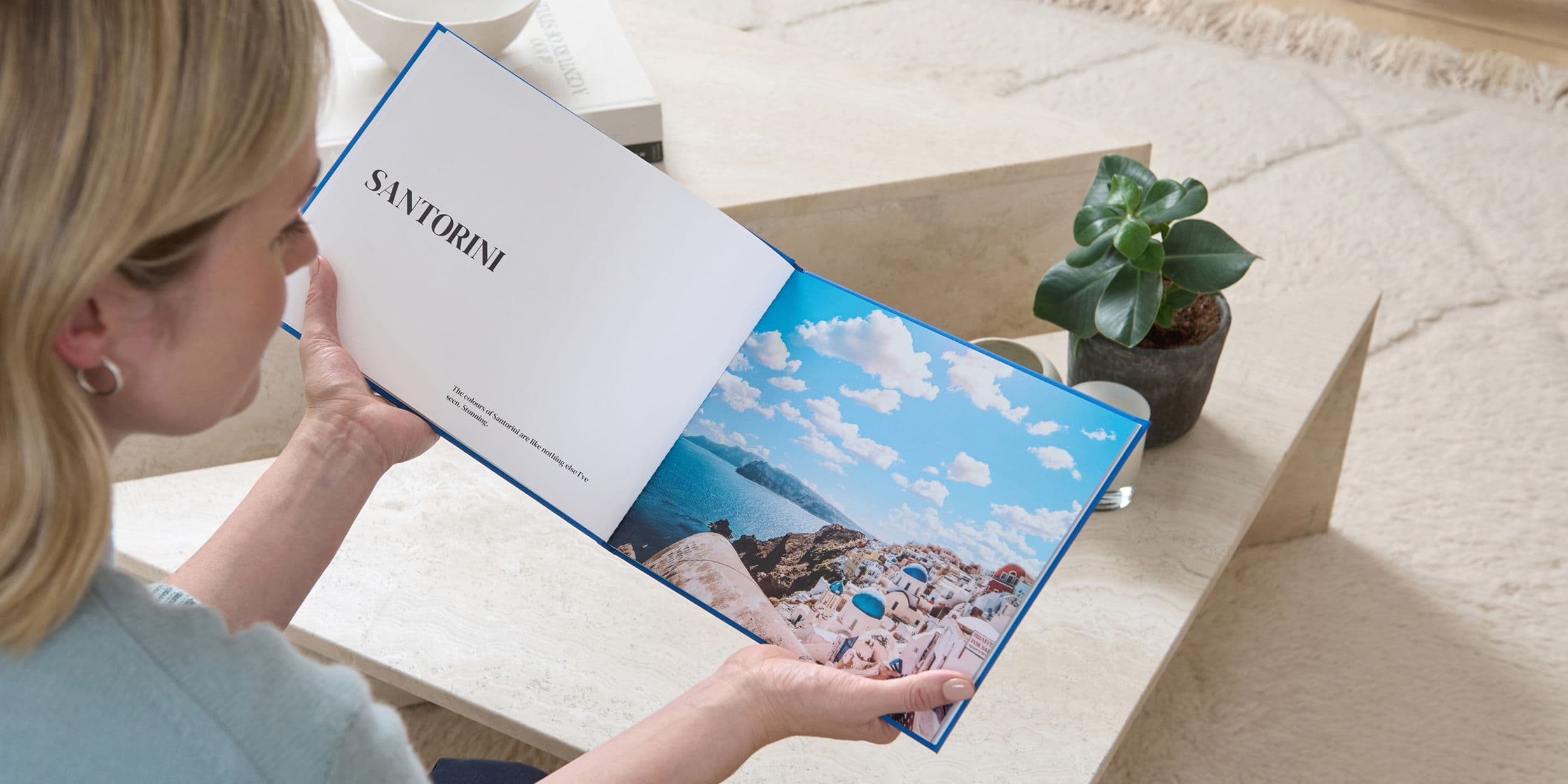 A person holds an open book with a page titled "Santorini" and a scenic photo of a coastline. A small plant is on a table nearby.