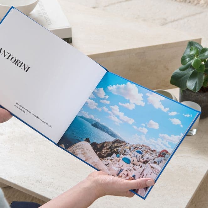 A person holds an open book with a page titled "Santorini" and a scenic photo of a coastline. A small plant is on a table nearby.