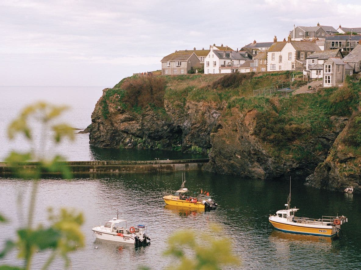 Coastal village with houses on a cliff, overlooking a calm sea with three small boats. Foreground features blurred greenery.