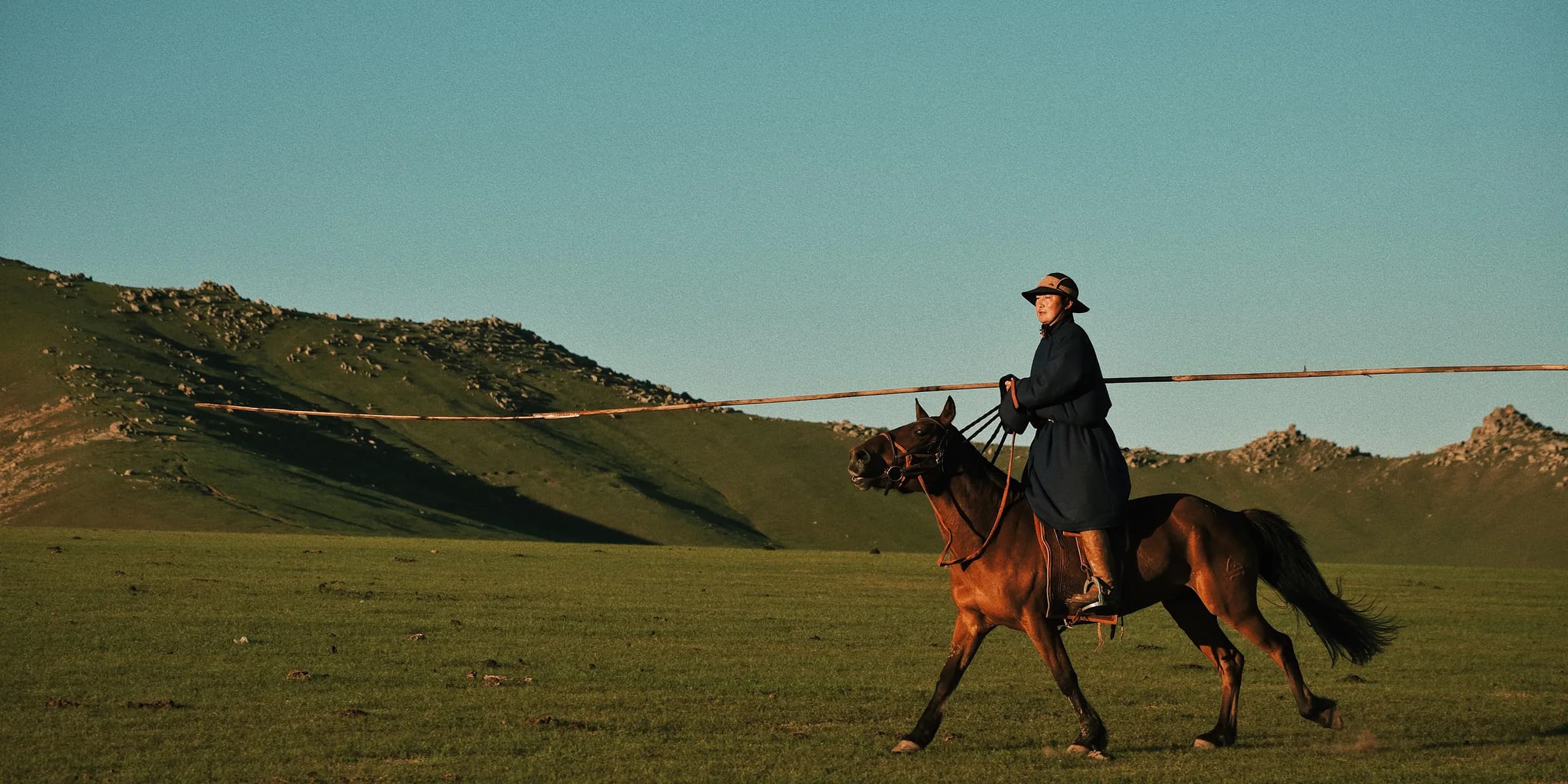 Person in a dark coat and hat rides a brown horse across a vast, grassy landscape with distant hills under a clear blue sky.