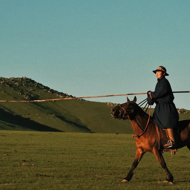 Person in a dark coat and hat rides a brown horse across a vast, grassy landscape with distant hills under a clear blue sky.