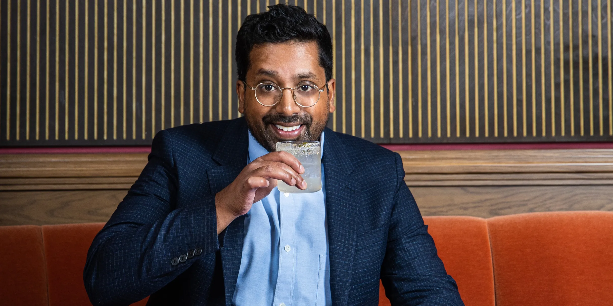 Man in a suit sitting on an orange bench, smiling, and sipping a drink from a glass with a salted rim.