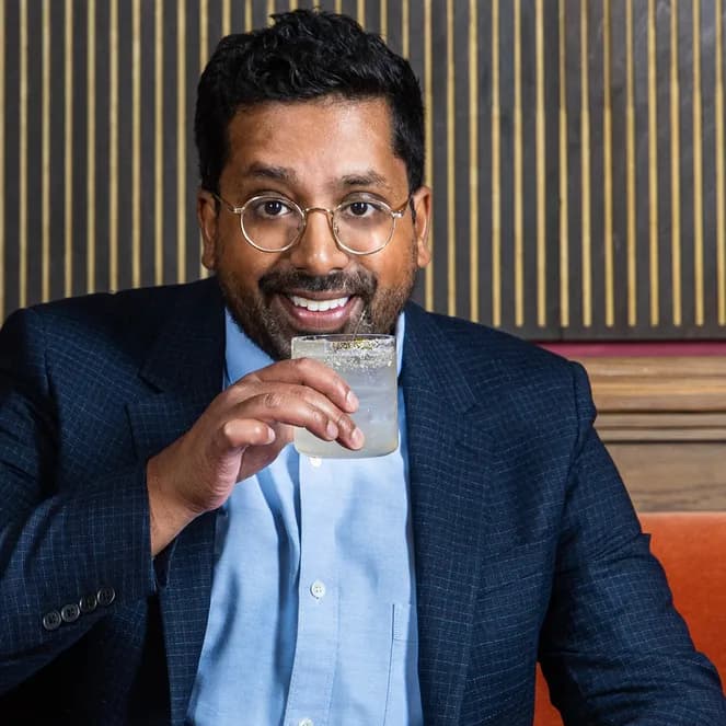 Man in a suit sitting on an orange bench, smiling, and sipping a drink from a glass with a salted rim.