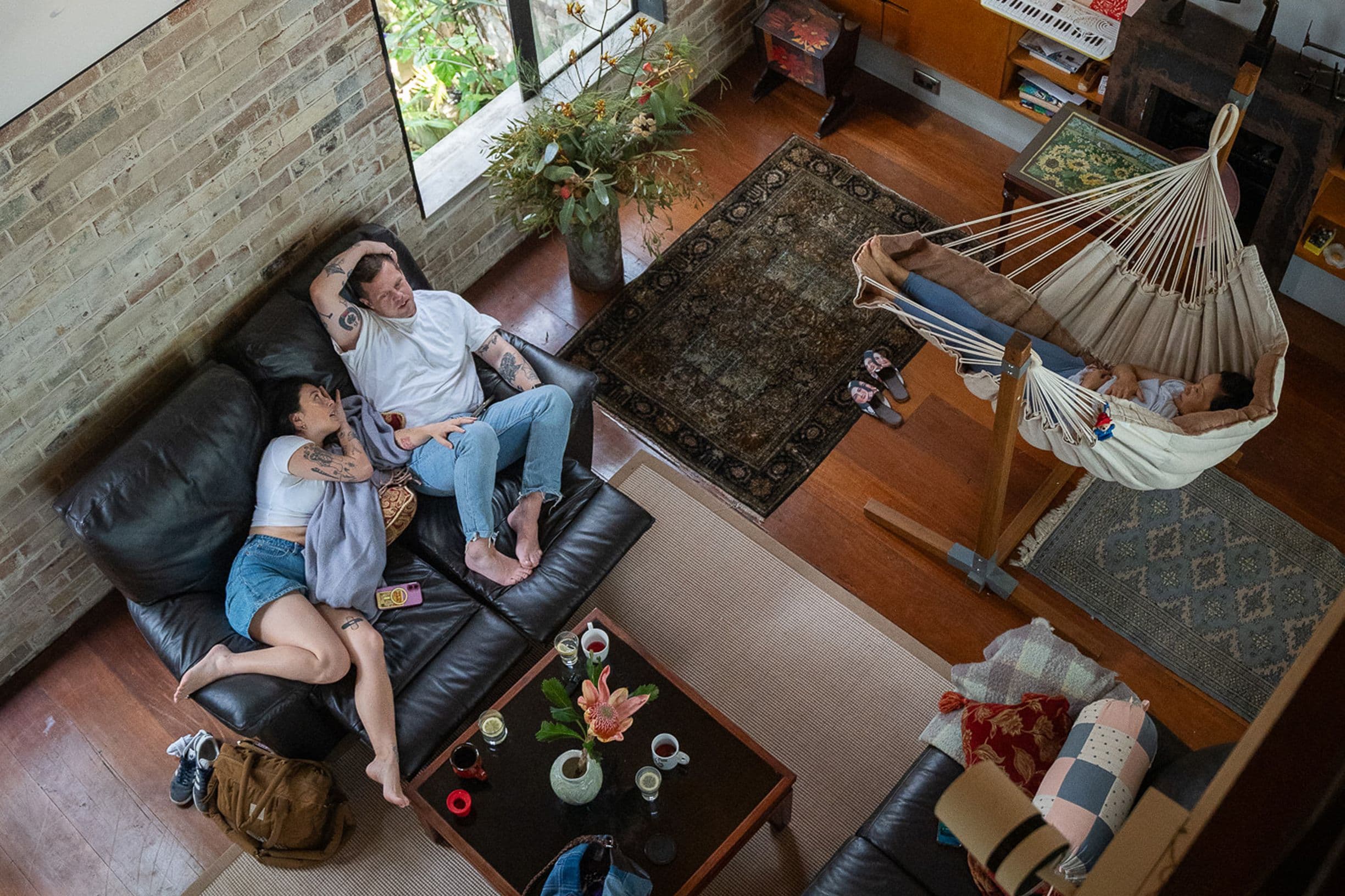 A couple relaxes on a sofa while a baby sleeps in a hammock in a cozy living room with wooden floors and a large window.