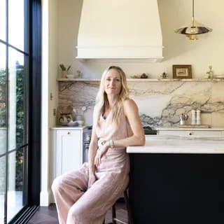 Woman in a light pink jumpsuit sits on a stool in a modern kitchen with marble backsplash, large window, and wooden floor.