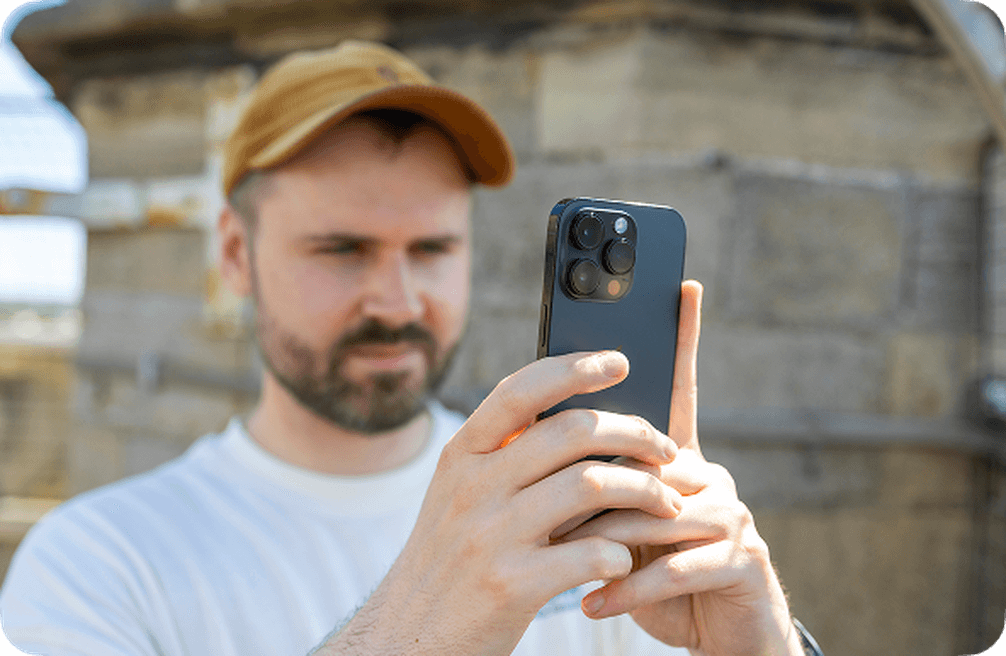 Dan, wearing a cap using an iPhone to take a photo outdoors, with a blurred background of a stone structure.