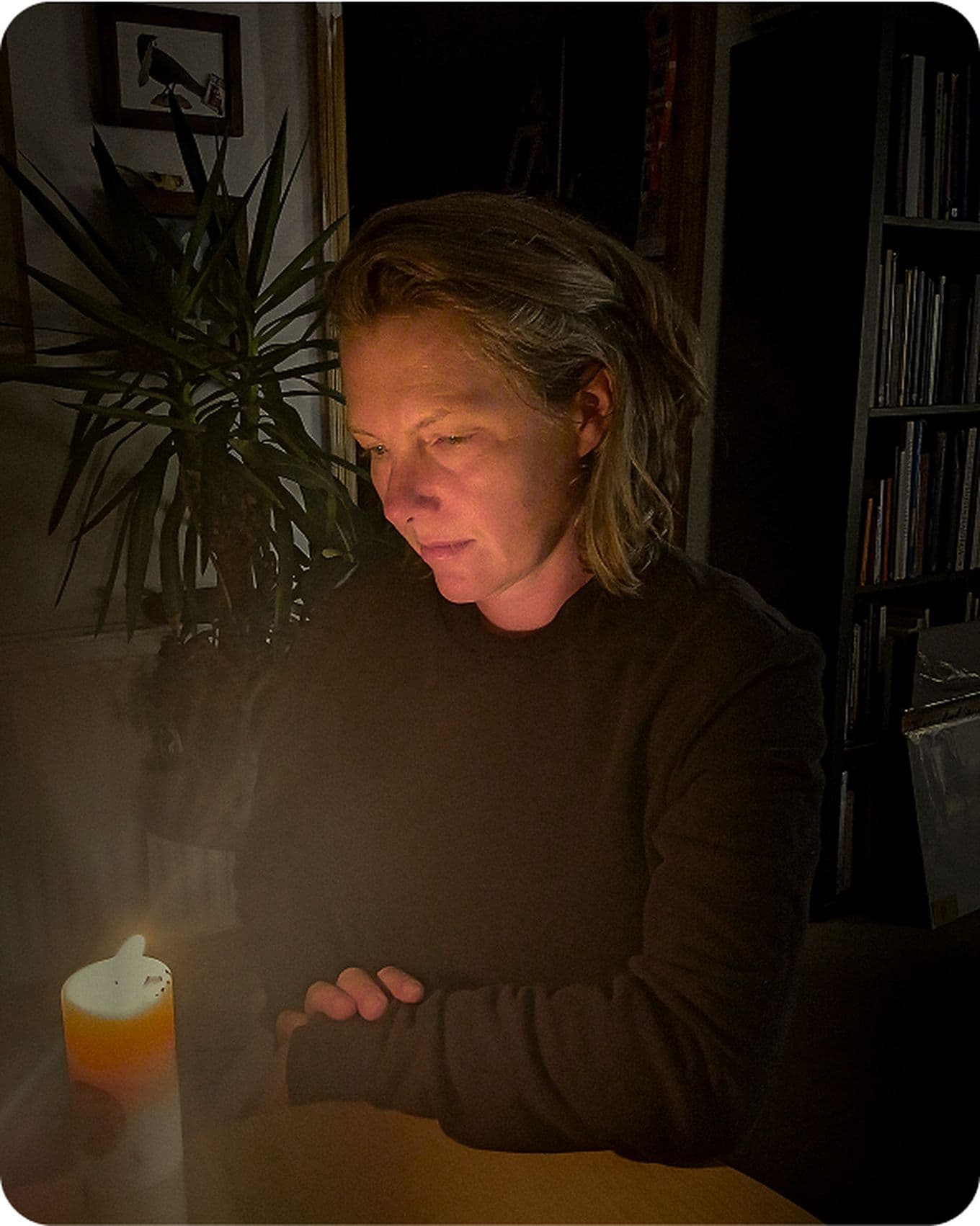 A person with short hair sits in dim light, gazing at a lit candle, with a plant and bookshelves in the background.