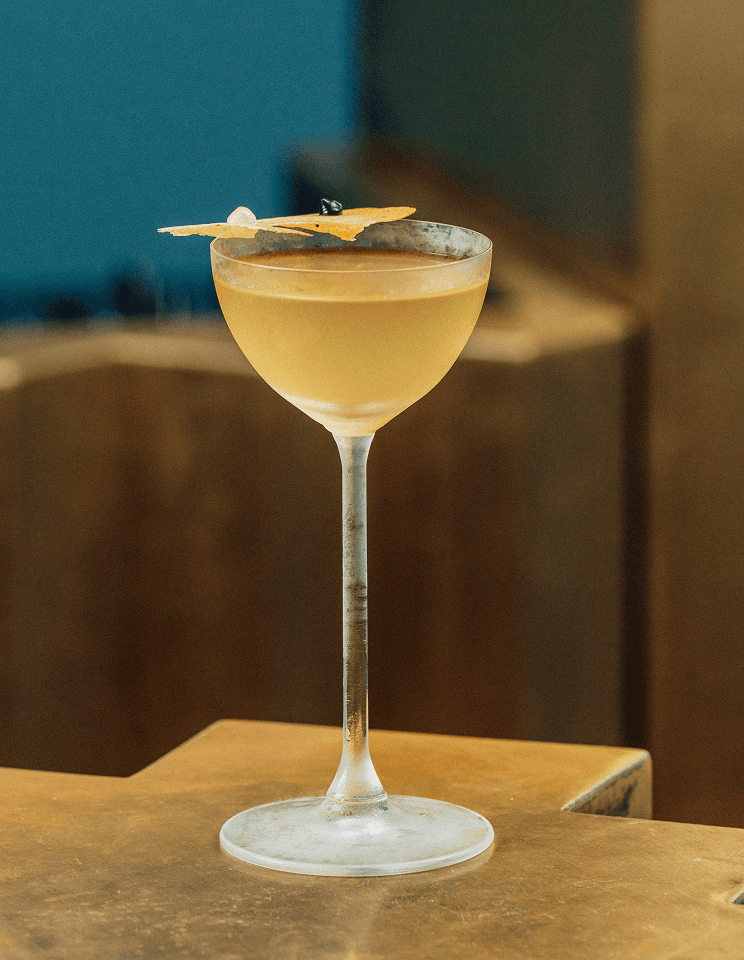 A frosted cocktail glass filled with a light-colored beverage, garnished with a slice of fruit and a small decorative item, placed on a wooden surface.