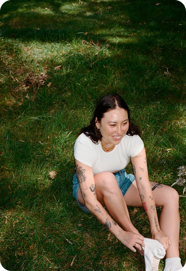 A person with tattoos sits on grass, wearing a white shirt and denim shorts, smiling while holding a white cloth.