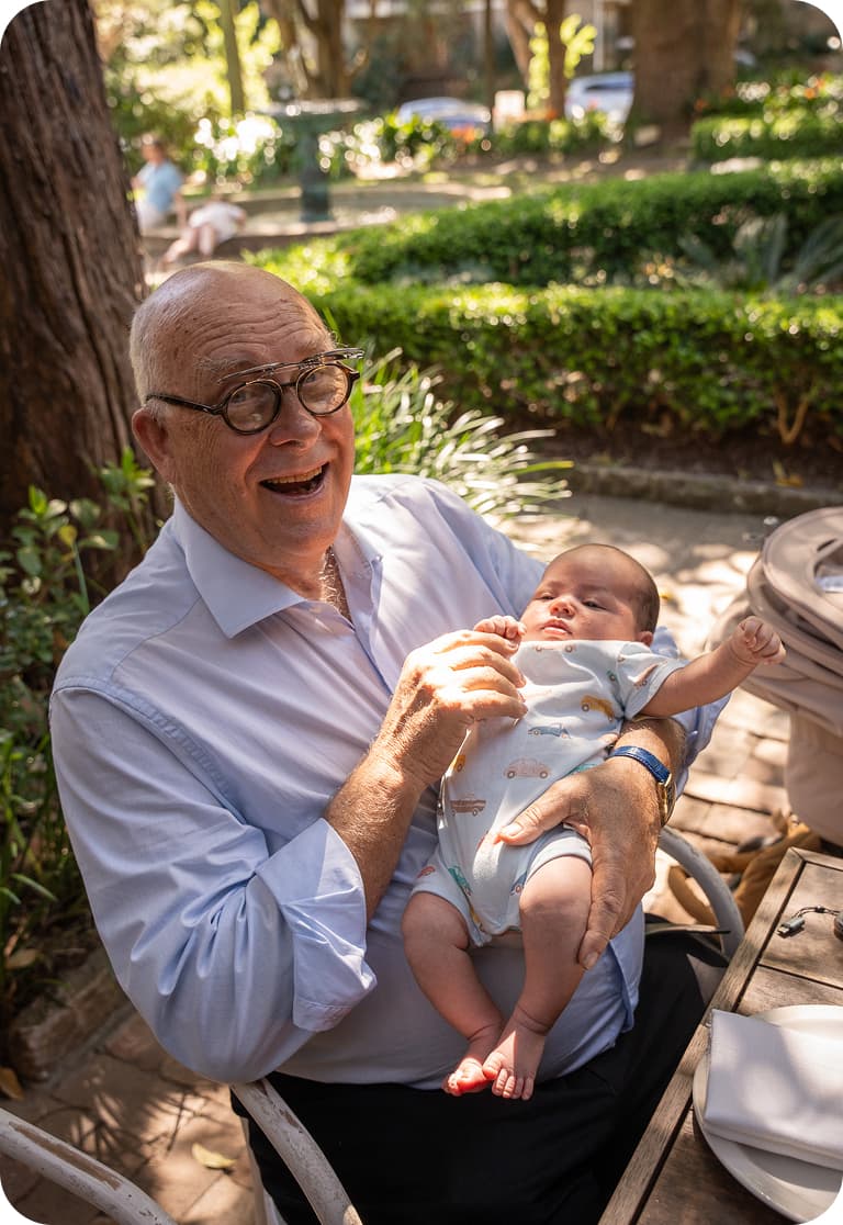 Elderly man joyfully holding a baby outdoors, surrounded by greenery and seated at a wooden table.