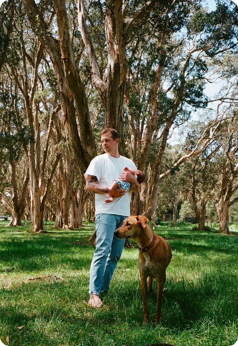 Man holding a baby stands in a sunlit forest with a large brown dog. Tall trees and green grass surround them.