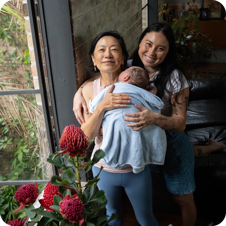 Two women smiling indoors, one holding a baby wrapped in a blue blanket. Red flowers are in the foreground near a window.