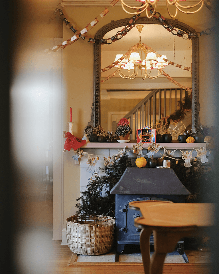 Cozy interior with a decorated mantelpiece, garland, mirror reflection, and a wood stove. Nearby, there's a wicker basket and wooden furniture.