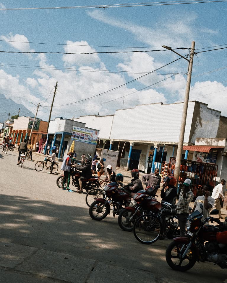Street scene with people on motorbikes and bicycles in front of shops on a sunny day, with a clear blue sky and scattered clouds.