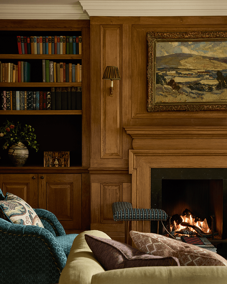 Cozy living room with a fireplace, artwork above, wooden bookshelves filled with books, patterned cushions on chairs, and a vase with flowers.