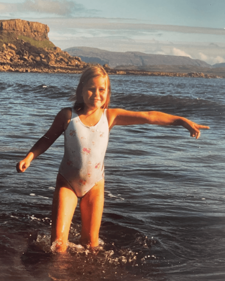 A young girl in a floral swimsuit walks through shallow water, smiling and pointing, with rocky cliffs and mountains in the background.