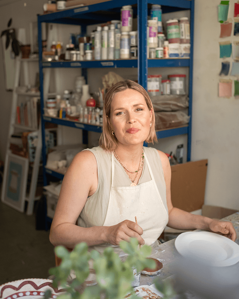 A woman in an apron sits at a table, holding a brush in a room filled with art supplies and shelves, with a relaxed expression.