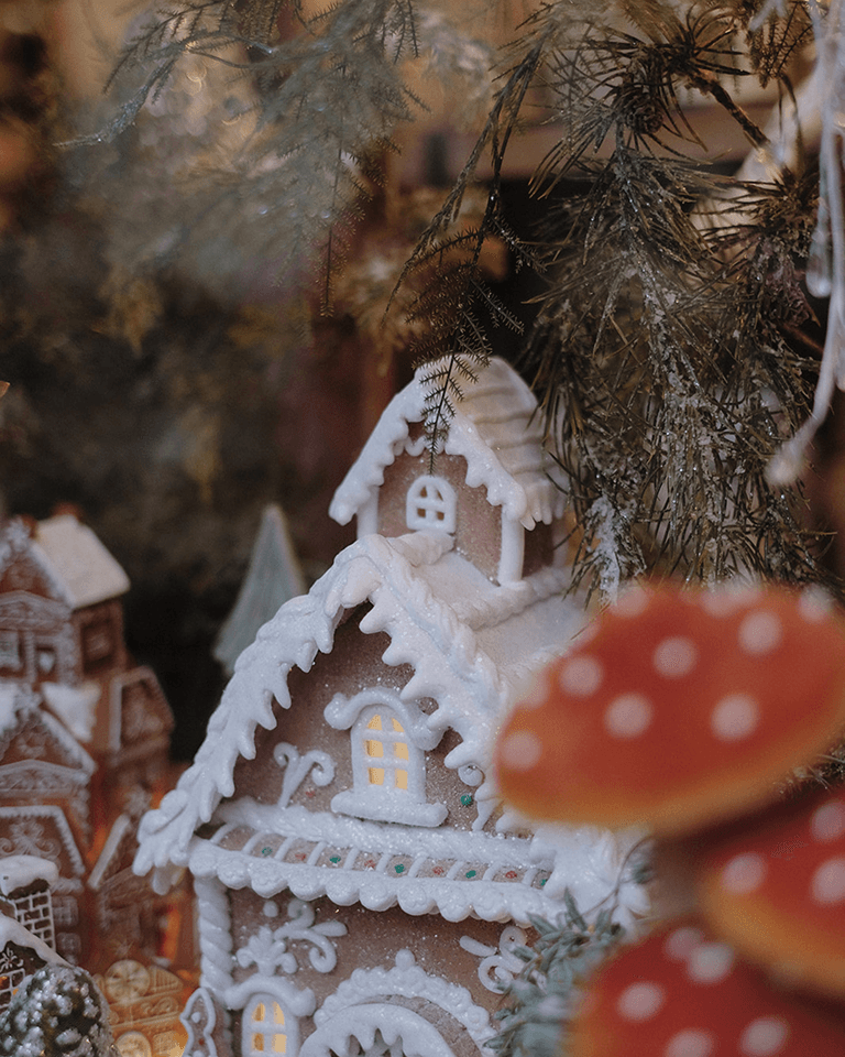 A gingerbread house with intricate icing details and lit windows, surrounded by festive greenery and blurred red mushrooms in the foreground.