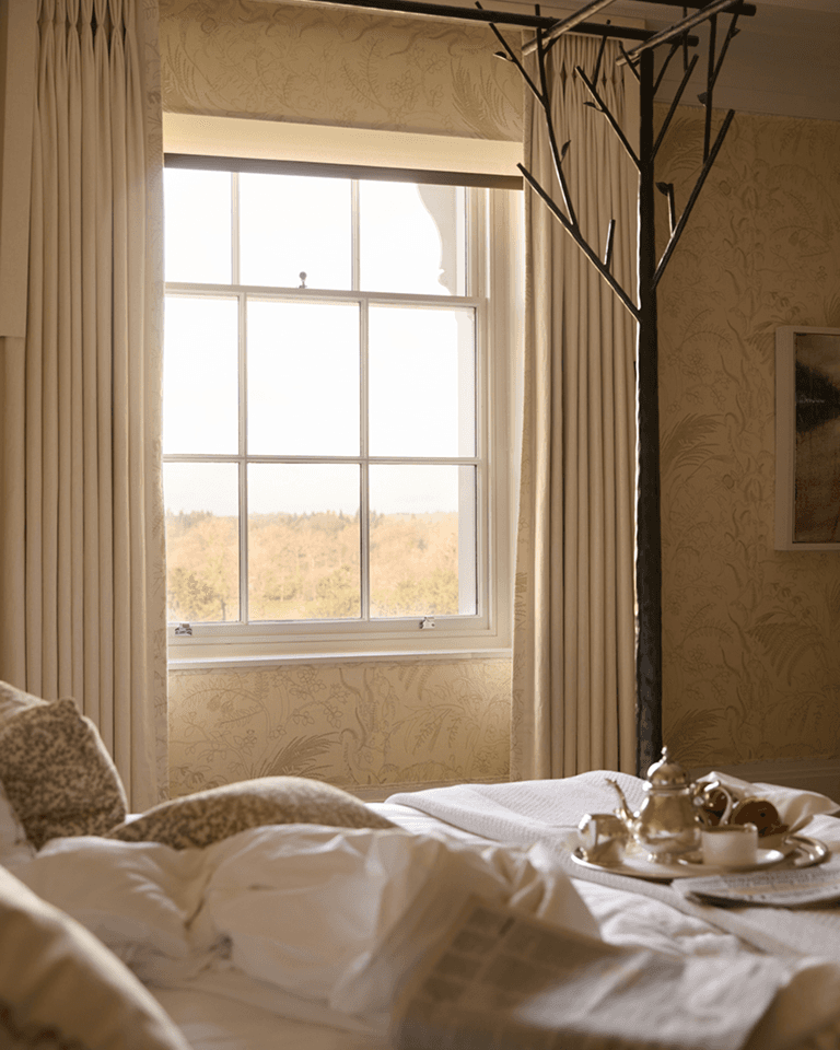 Cozy bedroom with a messy bed, a breakfast tray, and a tree-like coat rack. A window reveals a soft, scenic view outside.