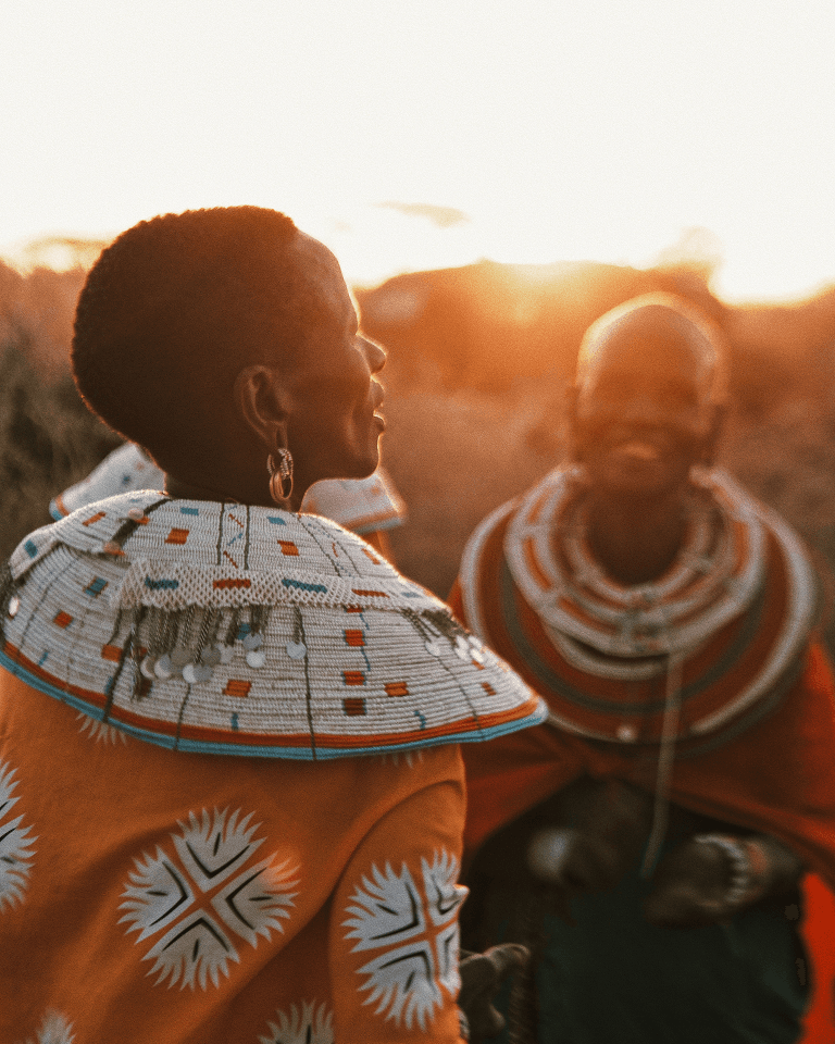 Two people wearing traditional, colorful beadwork and clothing, standing outdoors at sunset.
