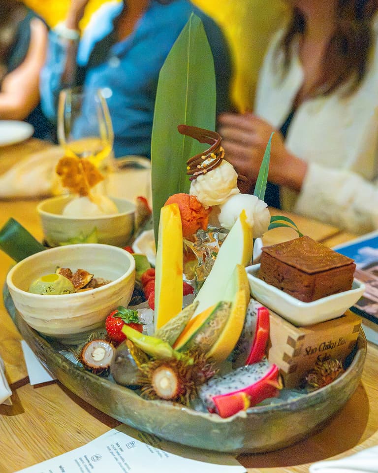 A platter of assorted tropical fruits, ice cream bowls, a chocolate dessert, and decorative leaves on a table with people in the background.