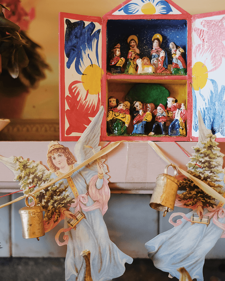 Colorful nativity scene with small figurines inside a painted wooden box. Angels and miniature Christmas trees decorate the foreground.