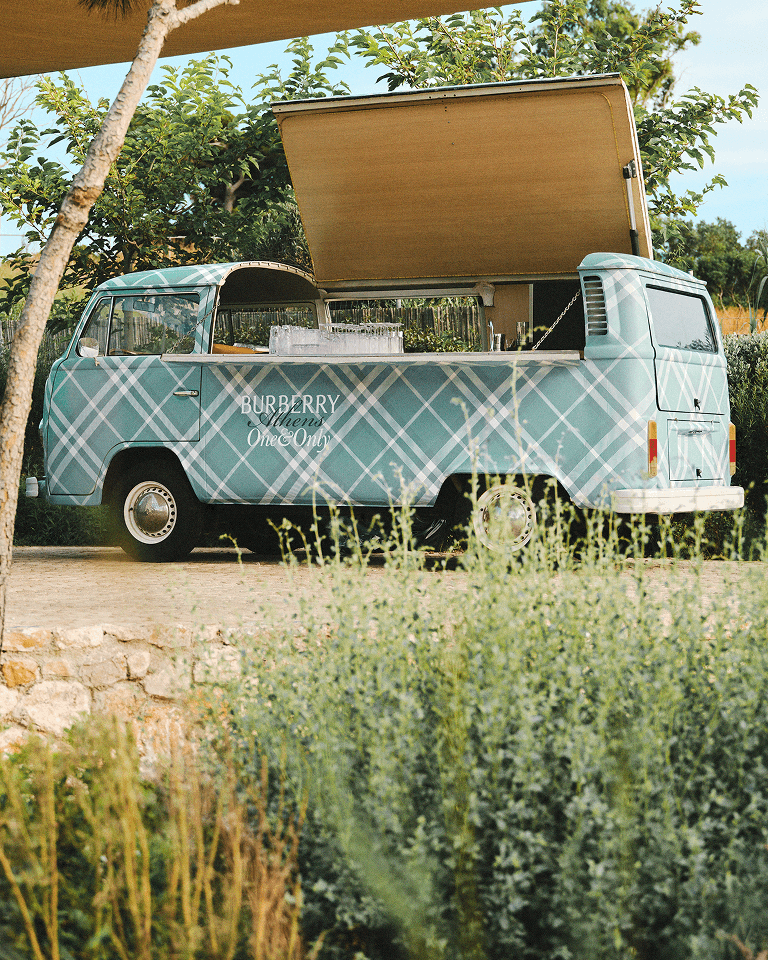 A vintage blue van with a plaid pattern, converted into an outdoor bar, parked among lush greenery with its side open.