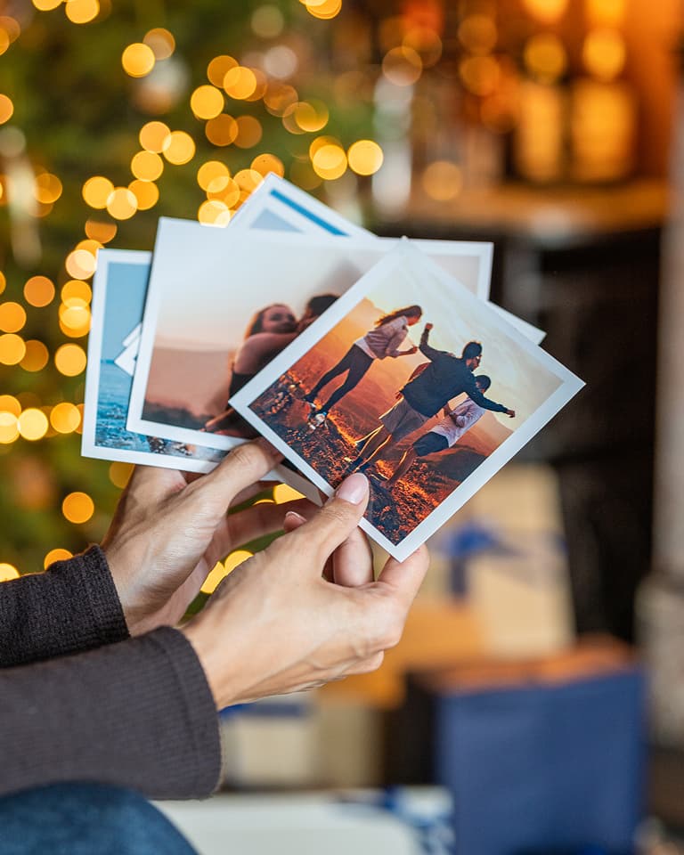 Hands holding a stack of printed photos, one showing joyful people outdoors. Background features blurred holiday lights and wrapped gifts.