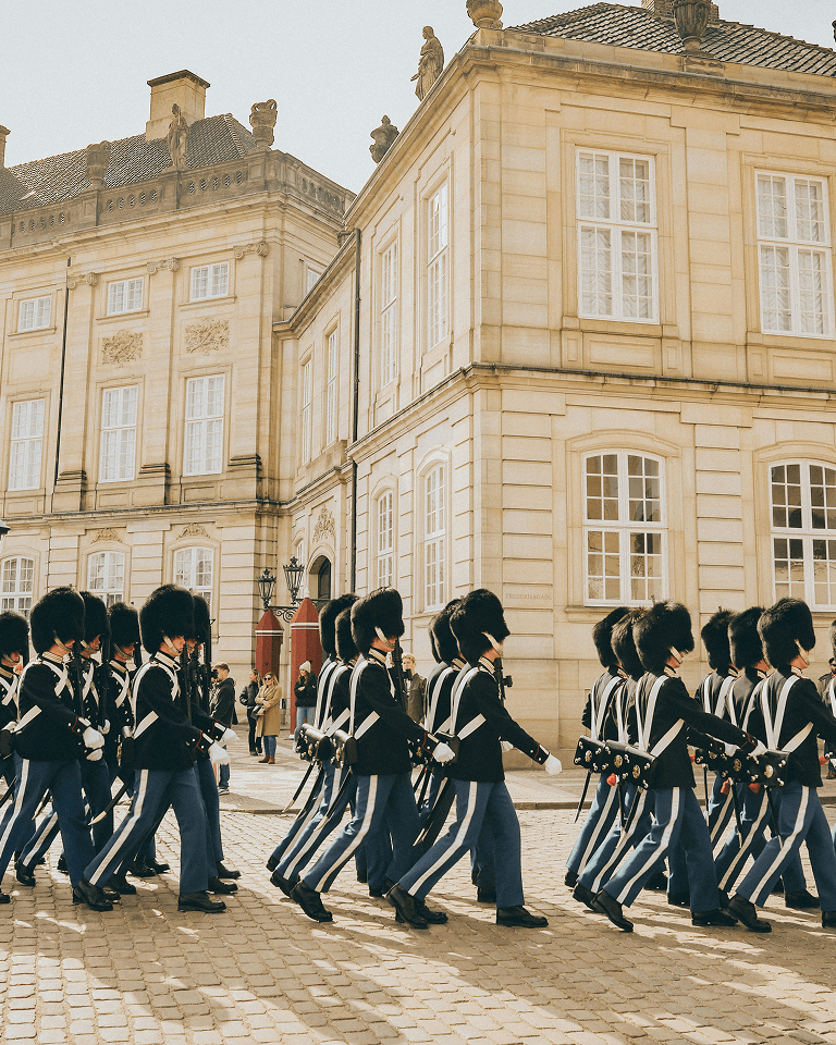 Guards in traditional uniforms and bearskin hats march in formation outside a historic beige stone building with large windows.